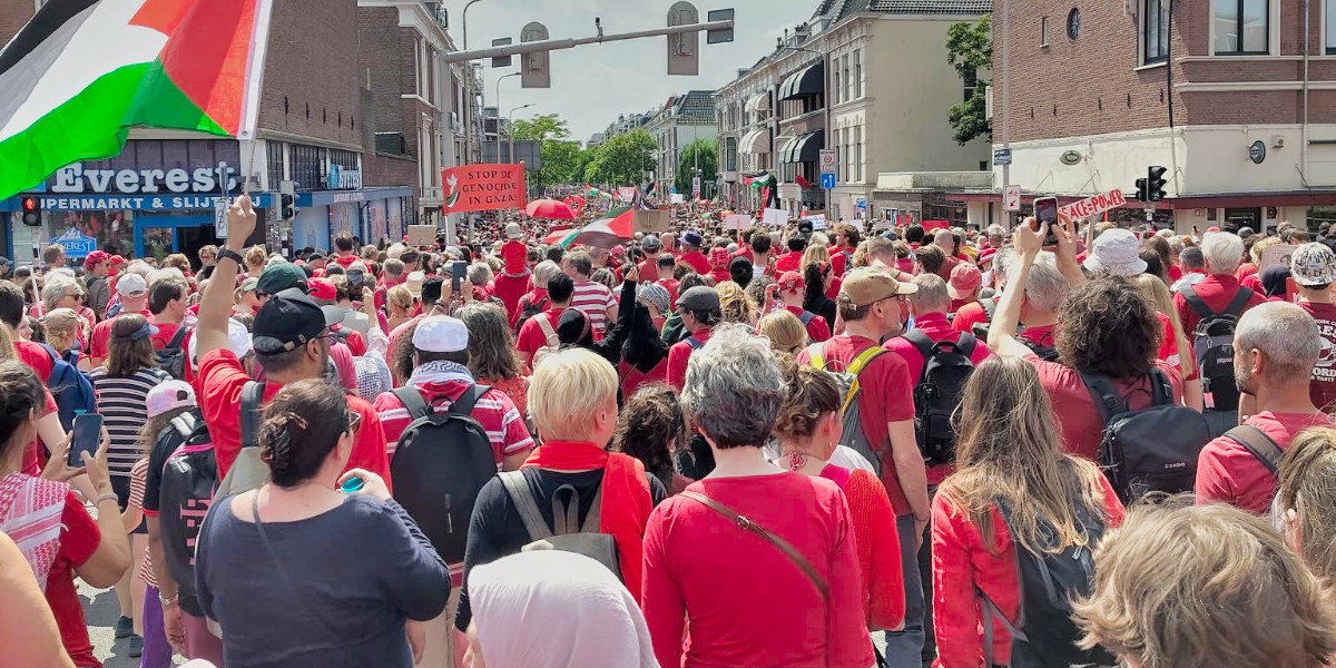 Massa van in het rood geklede mensen wringt zich door een smalle straat met een Palestijnse vlag erboven
