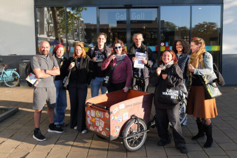 Groep BIJ1'ers voor een winkelcentrum, BIJ1-flyers in hun hand, rondom een bakfiets met actiestickers erop.