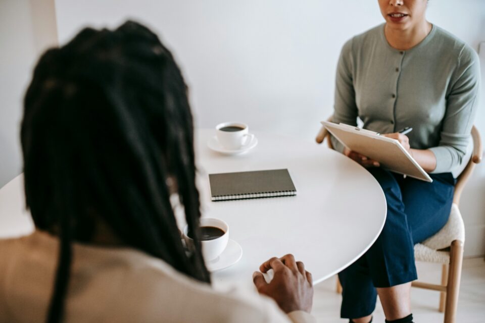 Twee personen in conversatie aan een tafel, waarbij er één aantekeningen maakt.